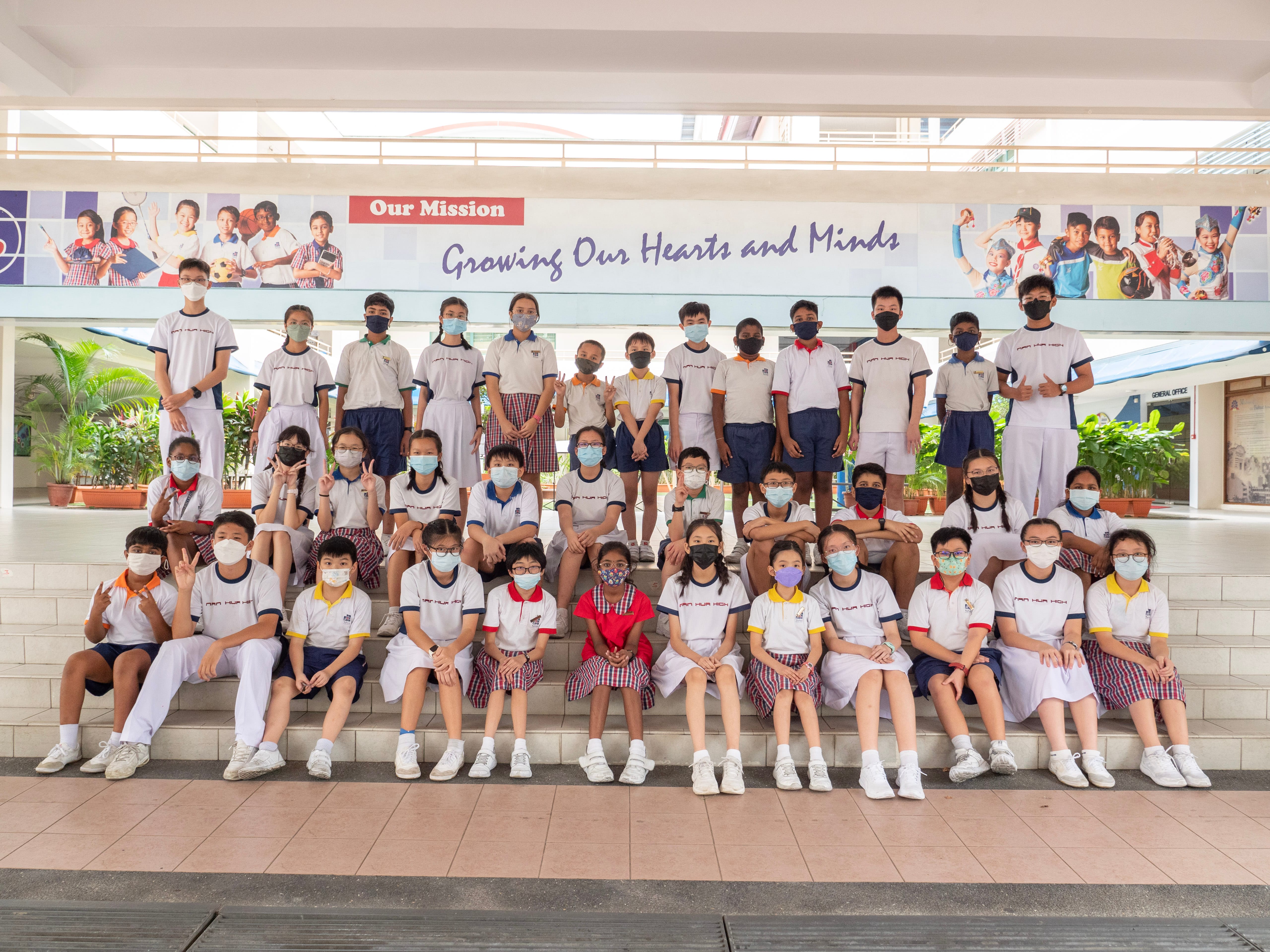A large group of masked school students in uniforms posed on steps beneath a banner reading Growing Our Hearts and Minds.